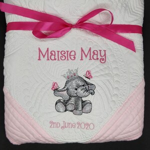 May include: A white and pink baby quilt with a pink ribbon tied around it. The quilt is embroidered with the name "Maisie May", a small elephant wearing a crown, and the date "2nd June 2020".