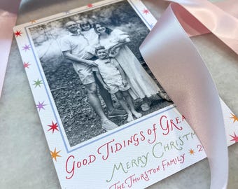 May include: A rectangular holiday card featuring a black and white family photograph surrounded by a border of colorful stars. The text reads Good Tidings of Great Joy, Merry Christmas, and The Thurston Family 2024. A light pink satin ribbon curves across the card.