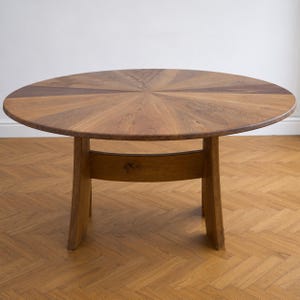May include: A round wooden dining table featuring a sunburst pattern on the tabletop surface. The table is constructed from light and dark brown wood segments and is supported by two curved legs connected by a horizontal wooden support beam.