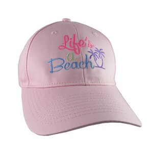May include: A pale pink baseball cap with a curved peak. The cap has the embroidered phrase "Life's a Beach" in pink, blue, and green, with a palm tree design. The cap is made of a soft material.