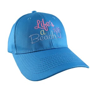 May include: A blue baseball cap featuring the embroidered text "Life's a Beach" in pink and green, and a palm tree design. The cap has a curved peak and a button on the crown.