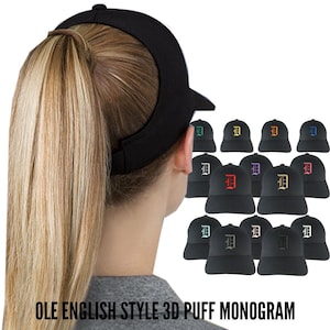 May include: Black visor with a curved peak, designed for a ponytail. The image also shows several black baseball caps, each embroidered with a capital "D" in various colours. Text at the bottom reads "OLE ENGLISH STYLE 3D PUFF MONOGRAM".