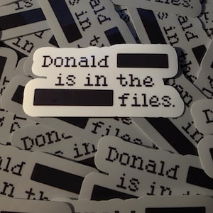 May include: A collection of white stickers with the text "Donald is in the files." printed in a pixelated font. The stickers have a black rectangle covering a portion of the text.