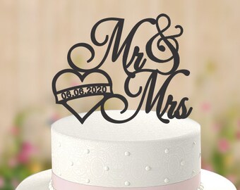 May include: A three-tiered white wedding cake with pink ribbon accents. A black cake topper reads "Mr & Mrs" with a heart shape that includes the date "06.06.2020". The cake has a delicate pattern.