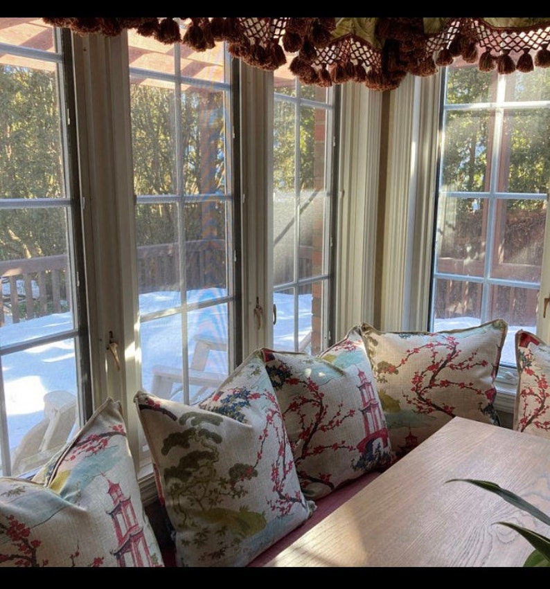 Pode incluir: Uma janela de ba&iacute;a com vista para um jardim nevado. A janela &eacute; decorada com uma cornija com borlas. Uma mesa de madeira com um conjunto de almofadas com padr&atilde;o floral fica em frente &agrave; janela.