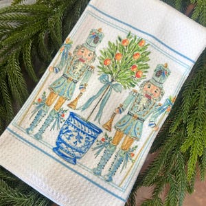 May include: White decorative kitchen towel featuring two nutcracker figures in light blue uniforms with gold accents. A blue and white planter and a tree with orange fruit are between the figures. The towel has a blue border and is decorated with a festive design.
