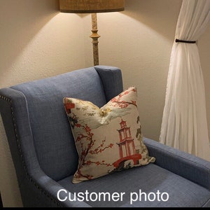 Pode incluir: Uma poltrona azul com um almofada estampada em branco e vermelho com um design de pagoda. A almofada tem um padr&atilde;o floral com flores cor-de-rosa. A poltrona est&aacute; em uma sala com uma l&acirc;mpada e cortinas brancas.