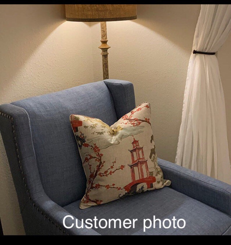 Pode incluir: Uma poltrona azul com um almofada estampada em branco e vermelho com um design de pagoda. A almofada tem um padr&atilde;o floral com flores cor-de-rosa. A poltrona est&aacute; em uma sala com uma l&acirc;mpada e cortinas brancas.
