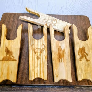 May include: A collection of wooden boot jacks with carved designs. The designs include a wolf, a bull skull, a cowboy on a horse, and a Bigfoot figure. The boot jacks are made of light-colored wood and are displayed on a dark wooden surface.