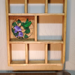 May include: Wooden wall shelf with multiple compartments. The shelf features a round, carved wooden accent with a green hummingbird and purple flowers. The shelf is made of light-colored wood and is designed for displaying small items.
