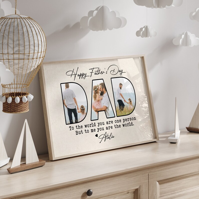 Op de afbeelding: Een ingelijste print met de woorden "Happy Father's Day" en "DAD" in een stijlvol lettertype. Het woord "DAD" bevat drie familiefoto's. De tekst hieronder luidt "To the world you are one person But to me you are the world." en is gesigneerd "Atalia."