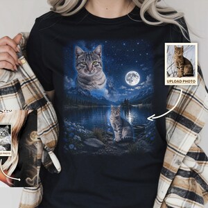 May include: A black t-shirt featuring a custom graphic print of a tabby cat in a night landscape with a full moon, mountains, and a lake. The design includes a floating cat head above the scene and a full-body cat sitting by the water's edge.