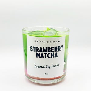 May include: A clear glass candle with a green wax top and a white label. The label reads "STRAWBERRY MATCHA Coconut Soy Candle 16oz". The candle has a white wick and is set against a white background.
