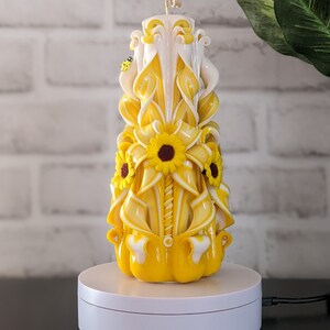 Könnte beinhalten: Eine gelbe und weiße geschnitzte Kerze mit einem Sonnenblumendesign. Die Kerze steht auf einem weißen Drehteller.
