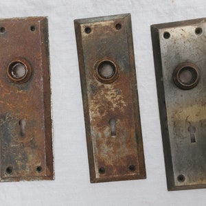 May include: Three antique door plates with keyholes and a round knob. The plates are made of metal and have a worn, rusty finish.