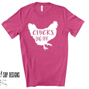 May include: A magenta t-shirt with a white graphic of a chicken and the text "CHICKS DIG ME". The shirt is a crew neck style and has short sleeves. The brand "SOP DESIGNS" is printed in black on the bottom left.