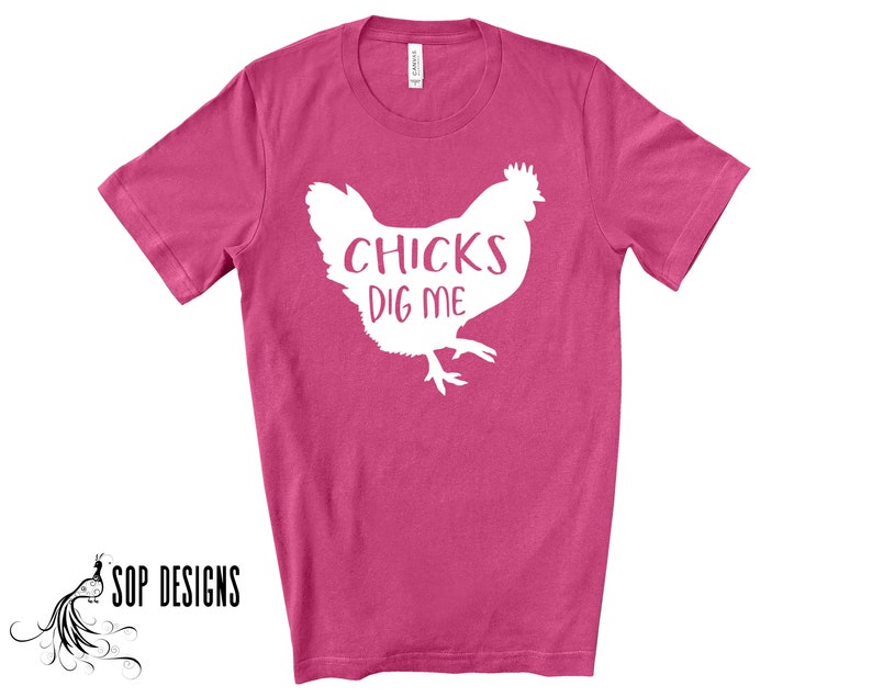 May include: A magenta t-shirt with a white graphic of a chicken and the text "CHICKS DIG ME". The shirt is a crew neck style and has short sleeves. The brand "SOP DESIGNS" is printed in black on the bottom left.