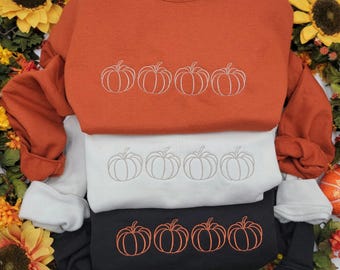 May include: Three sweatshirts in shades of orange, white, and black, each embroidered with four pumpkin outlines. The sweatshirts are folded and stacked. The background features sunflowers and autumn foliage.