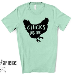 May include: A light green t-shirt with a black silhouette of a chicken and the words "CHICKS DIG ME" printed on the front. The shirt is made of a soft material and has short sleeves. The brand "SOP DESIGNS" is printed on the bottom left.