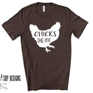 May include: A dark brown t-shirt with a white graphic of a chicken and the text "CHICKS DIG ME". The shirt has short sleeves and a crew neck. The brand "SOP DESIGNS" is printed in black on the bottom left.