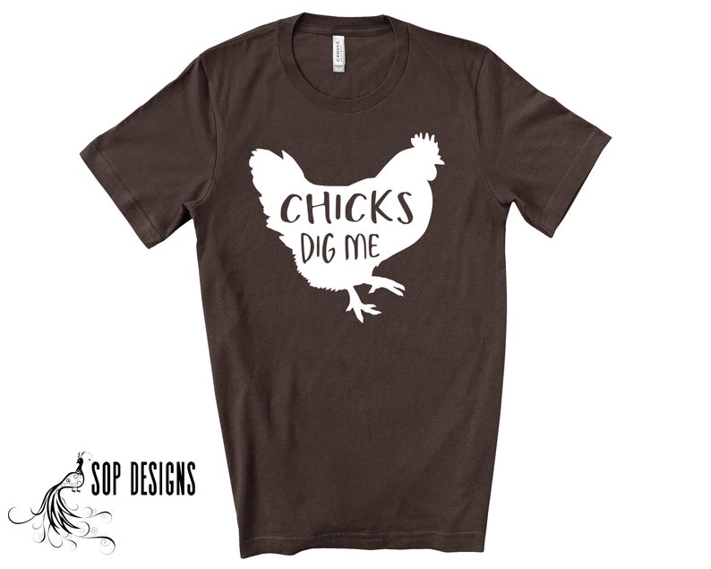 May include: A dark brown t-shirt with a white graphic of a chicken and the text "CHICKS DIG ME". The shirt has short sleeves and a crew neck. The brand "SOP DESIGNS" is printed in black on the bottom left.
