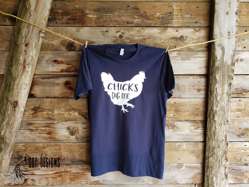 May include: Navy blue t-shirt with a white graphic of a chicken and the text "CHICKS DIG ME." The shirt is hanging on a clothesline against a rustic wooden background.