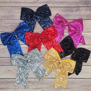 Puede incluir: Siete lazos para el pelo con lentejuelas en varios colores, incluyendo azul, rojo, rosa, negro, plateado, dorado y amarillo. Los lazos están dispuestos en un círculo sobre una superficie de madera.