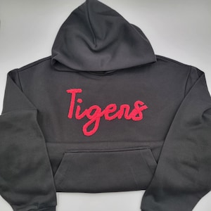 May include: A black hooded sweatshirt with the word "Tigers" in red script across the chest. The sweatshirt has a front pocket and a drawstring hood. The fabric appears to be a soft, comfortable material.