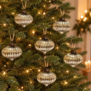May include: Six silver-toned, ribbed, onion-shaped glass ornaments hanging from a green pine tree. Each ornament features a metallic, reflective finish with vertical ridges and a small metal cap at the top, suspended by a simple brown twine loop.