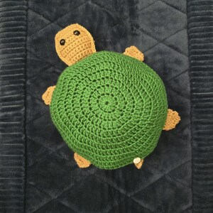 May include: A crochet turtle toy featuring a large, circular, dark green shell with a textured stitch pattern. The head and four small flippers are crafted in a light tan yarn. The turtle has two small, round, black plastic eyes on its head.