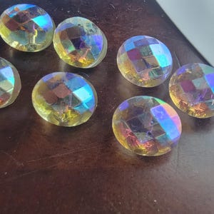 May include: Six round, faceted buttons with a clear, iridescent finish that reflects shades of blue, purple, and yellow. Each button features a geometric, diamond-cut pattern on the surface, creating a shimmering, multi-colored effect.