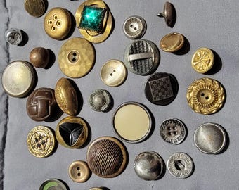 May include: A collection of vintage buttons in various colors, shapes, and sizes. The buttons are made of metal, plastic, and fabric. Some buttons have intricate designs, while others are plain. The buttons are arranged on a gray surface.