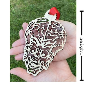 May include: A wooden ornament shaped like a skull with a detailed design. The ornament is painted in a light brown color and has a red ribbon attached to the top. The ornament is approximately 4 inches tall.