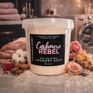 Può includere: Un contenitore di plastica trasparente con sapone per bucato con la scritta "Cashmere Rebel" su un'etichetta nera. L'etichetta recita anche "Oxy-Boosted Laundry Soap" e "32 oz." Il contenitore è circondato da fiori rosa, mandorle e altri elementi decorativi.