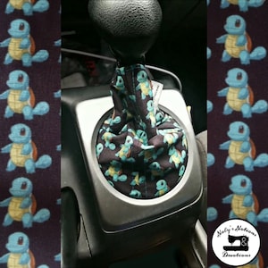 Puede incluir: Funda de palanca de cambios de tela negra con un patrón repetido de un personaje de tortuga de dibujos animados azul con vientre amarillo. La funda está instalada en la palanca de cambios de un coche. El fondo es una tela morada oscura con el mismo patrón de tortuga.