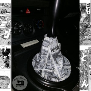 May include: A black gear shift knob with a fabric boot featuring a black and white manga print. The fabric displays various scenes of cars and Japanese text. The gear shift is installed in a car interior with black plastic dashboard controls.