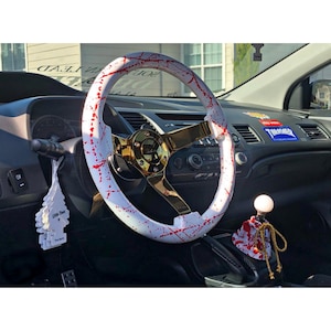 May include: White steering wheel with red splatters and a gold centre, and a white gear shift knob with red splatters in a car interior.
