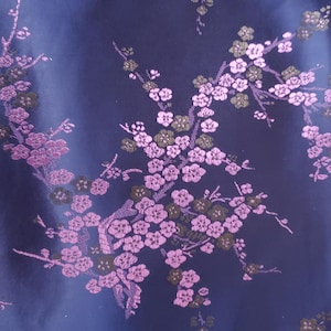 May include: Close-up of a navy blue fabric with a floral pattern. The design features branches with pink and olive-green blossoms. The fabric has a subtle sheen, suggesting a satin or silk material.