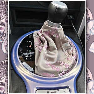 以下が含まれることがあります： 車のシフトノブのクローズアップ。花柄のファブリックカバーが付いています。シフトノブは、ギアポジション（P、R、N、M、D）を示す黒いマークが付いたシルバーリングで囲まれています。リングの下には「SPORT」の文字が見えます。