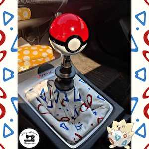 Puede incluir: Una palanca de cambios de Pokeball roja y blanca con una base negra. La palanca está unida a una funda de palanca de cambios blanca con un patrón geométrico rojo, azul y blanco. La funda está etiquetada con "WRX".