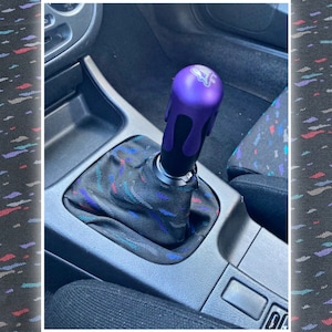 May include: Purple and black car gear shift knob with a drip design. The knob is installed in a car with a black and blue patterned gear shift boot.
