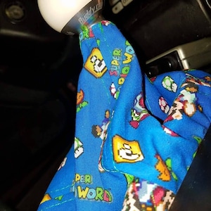 Puede incluir: Una funda para pomo de cambio de marchas azul con un patrón de Super Mario World. La funda tiene un pomo de cambio de marchas de bola blanca con el texto "Buddy Club" impreso en él.