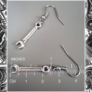 May include: Silver-colored wrench earrings. The earrings are shaped like miniature wrenches, with a hook at the top for the ear. The image shows the earrings against a gray background with a ruler in inches.