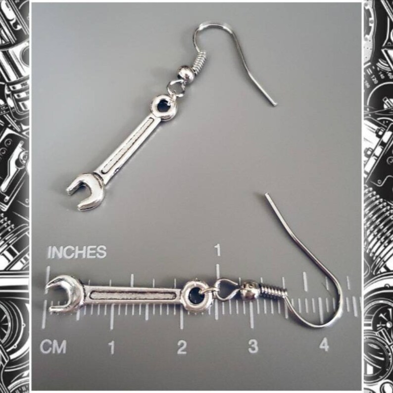 May include: Silver-colored wrench earrings. The earrings are shaped like miniature wrenches, with a hook at the top for the ear. The image shows the earrings against a gray background with a ruler in inches.