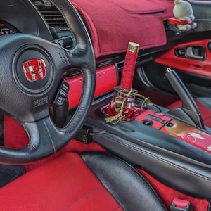 Puede incluir: El interior de un Honda S2000 rojo con un volante de cuero negro, asientos de cuero rojo y una palanca de cambios personalizada roja y dorada. El coche tiene una consola central de cuero negro con una funda de palanca de cambios roja y dorada.