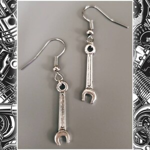 May include: A pair of silver-colored wrench earrings. Each earring features a small wrench charm suspended from a hook. The wrenches have a detailed, realistic design, with a small circular accent at the top and bottom.