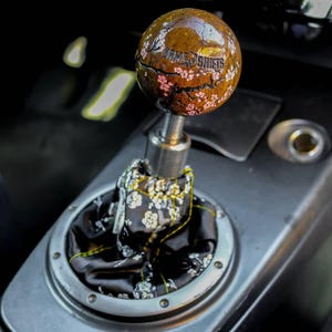 May include: A wooden gear shift knob with a cracked surface and pink cherry blossom design. The knob is engraved with the text "WRM SHIFTS". The knob is attached to a black gear shift boot with a white floral pattern.