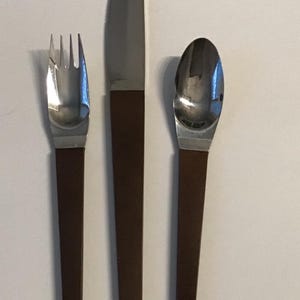 May include: A set of stainless steel cutlery, including a fork, knife, and spoon. The working ends are silver, and the handles are dark brown. The knife has a rounded tip. The set is displayed on a white surface.