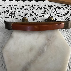 May include: A drawer pull featuring a curved, smooth, amber-colored insert set within a dark, weathered metal frame. The hardware includes two circular mounting posts on the back, showcasing a contrast between the polished resin and the aged metal finish.