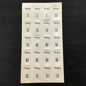 Pode incluir: Uma folha de etiquetas brancas com a marca "Bailey" impressa no topo de cada etiqueta. Cada etiqueta tem um número preto que indica tamanhos como 7, 7 1/2, 7 5/8, 7 1/8, 7 3/8, 6 7/8, 6 3/4, 6 5/8 e 7 3/8.